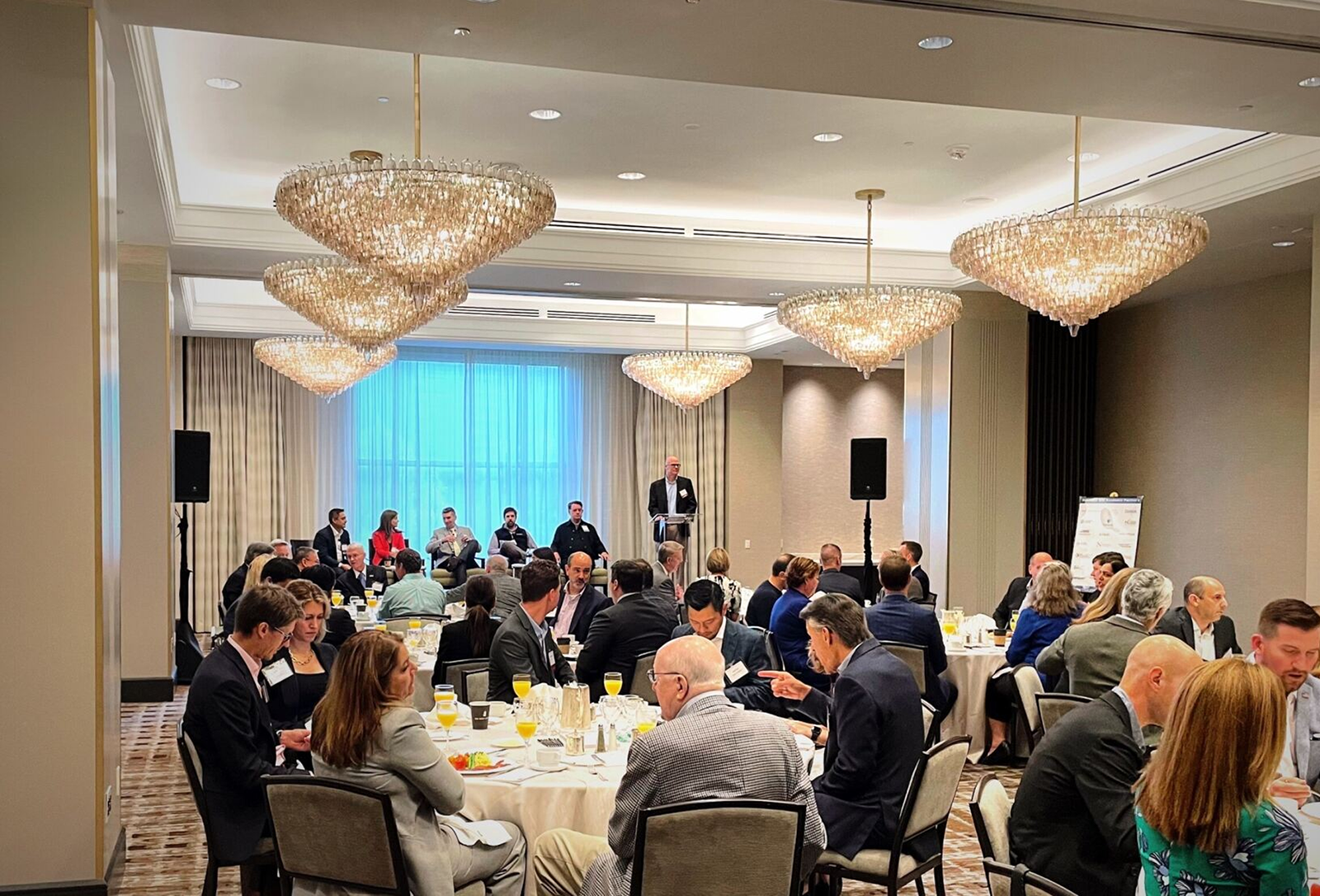 Breakfast reception around circular tables, with panelists in the back of the photo on a stage.